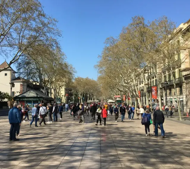 Tłum ludzi spaceruje po słonecznej La Rambla w Barcelonie, otoczonej drzewami i zabytkowymi budynkami.