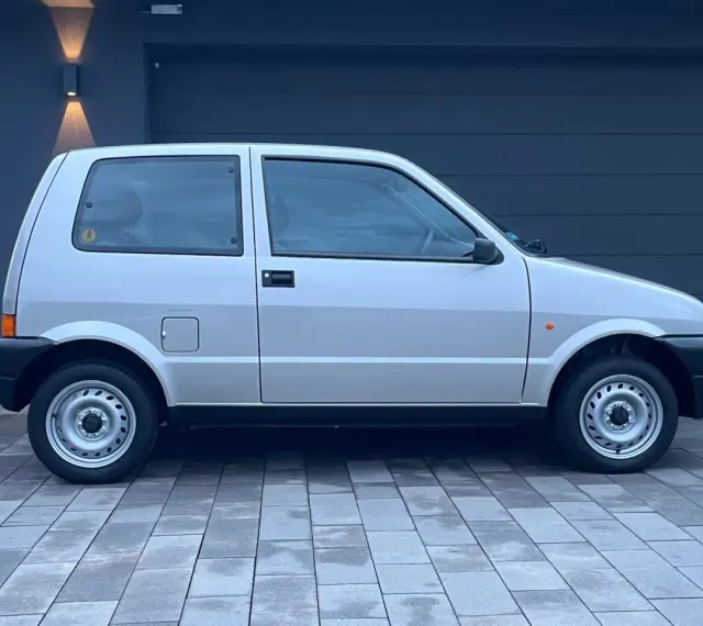 Srebrny Fiat Cinquecento na tle ciemnego garażu, z charakterystycznymi felgami i czarnymi zderzakami.