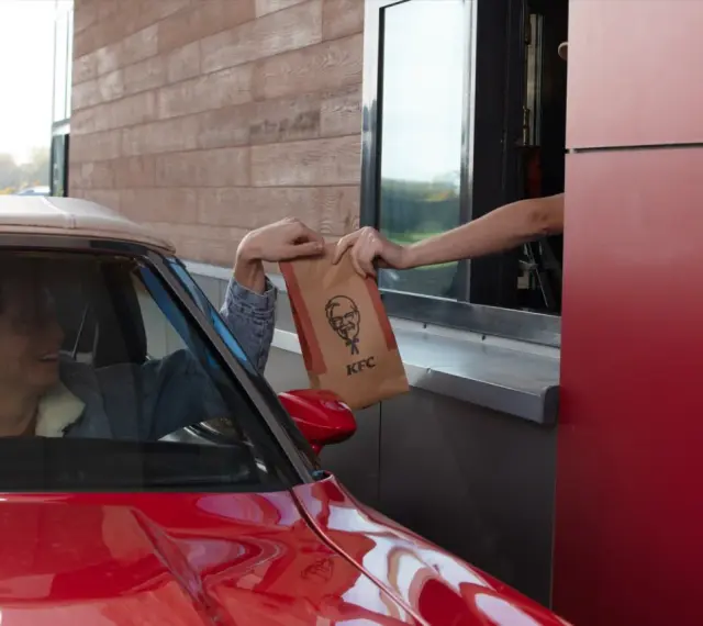Mężczyzna w czerwonym aucie odbiera z okienka KFC torbę z jedzeniem. Wygląda na zadowolonego, że otrzymał swój mega pocket kcal.