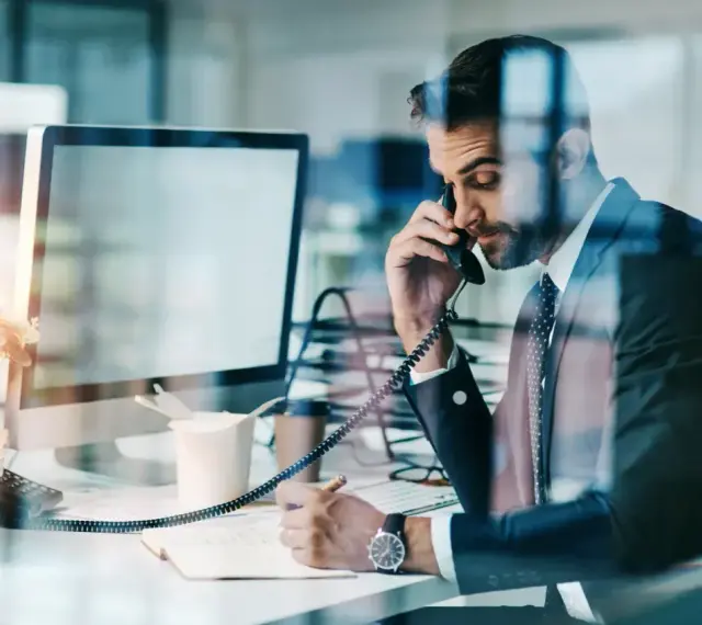 Biznesmen w garniturze rozmawia przez telefon, notując w zeszycie. Nawet w obliczu siły wyższej, praca wre.