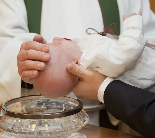 Chrzest dziecka w białej szatce. Woda święcona spływa po główce niemowlęcia do ozdobnej misy.