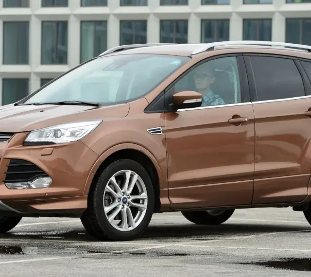 Brązowy Ford Kuga 2019 z kierowcą w okularach, zaparkowany na mokrym parkingu przed nowoczesnym budynkiem.