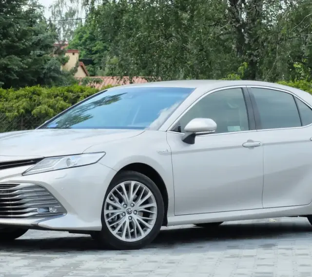 Srebrna Toyota Camry 2.5 Hybrid stoi na wybrukowanym podjeździe, otoczona zielenią.