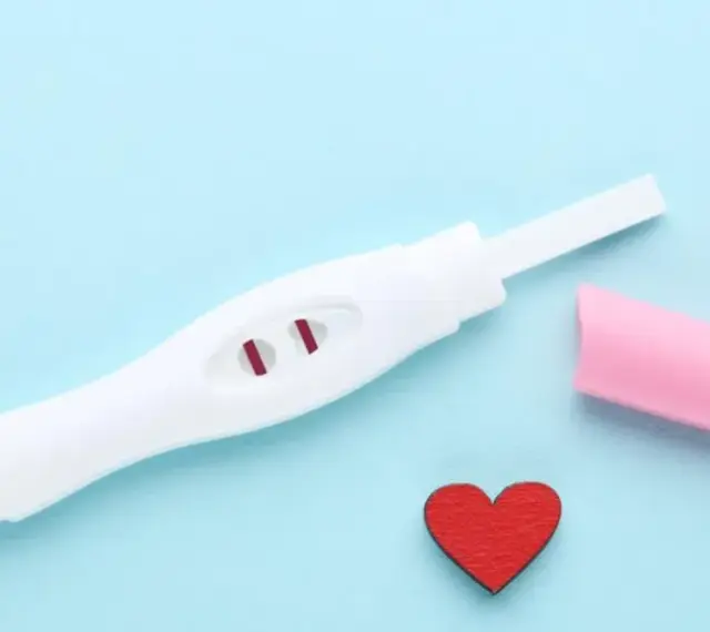 Biały test ciążowy z dwoma czerwonymi paskami, różowa nakładka i czerwone serduszko na niebieskim tle.
