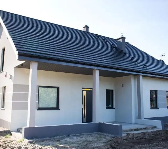 Nowy dom z szarą dachówką i jasną elewacją, ozdobioną subtelnymi, poziomymi pasami. Trwają prace nad malowaniem elewacji, nadając budynkowi nowoczesny wygląd.