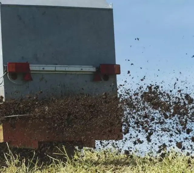 Rozrzutnik rozsypuje komunalne osady ściekowe na polu. Materiał jest wyrzucany w powietrze, tworząc brązową chmurę.
