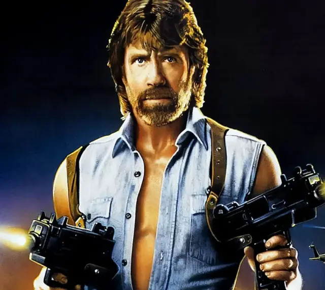 Chuck Norris z karabinami w akcji, jak w jego najlepszych filmach. W tle helikoptery i budynek.