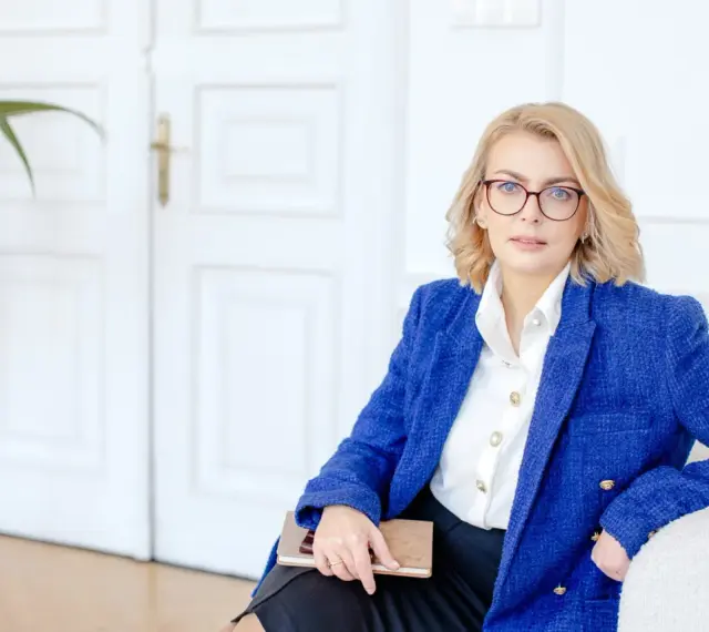 Kobieta w okularach i niebieskiej marynarce trzyma notatnik. Planuje dofinansowanie na otwarcie firmy.