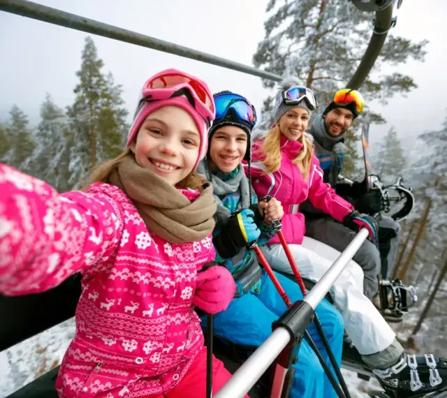 Rodzina na wyciągu narciarskim, robiąca selfie. Dziewczynka z przodu uśmiecha się szeroko. To ich wymarzone wczasy pod gruszą, ale zimowe!