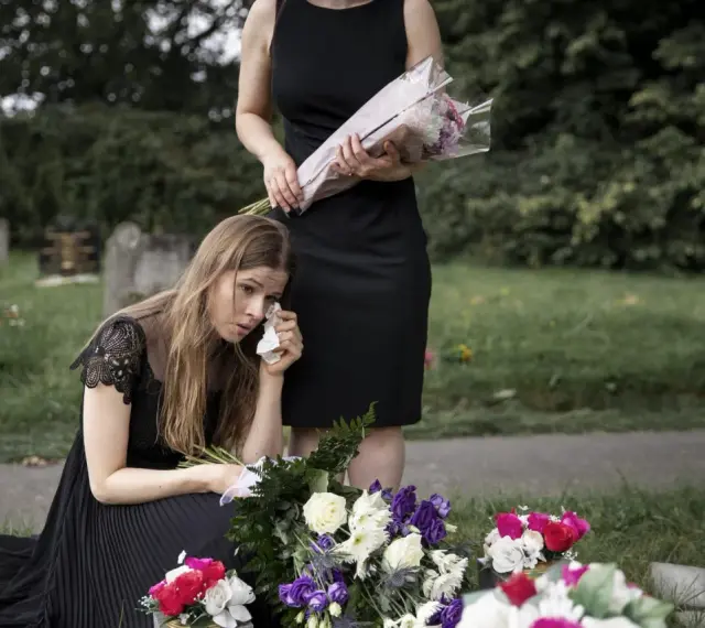Smutna kobieta w czarnej sukience klęczy przy grobie, ocierając łzy. Obok stoi druga kobieta z bukietem kwiatów. To obraz żałoby, trudny urlop macierzyński po poronieniu.