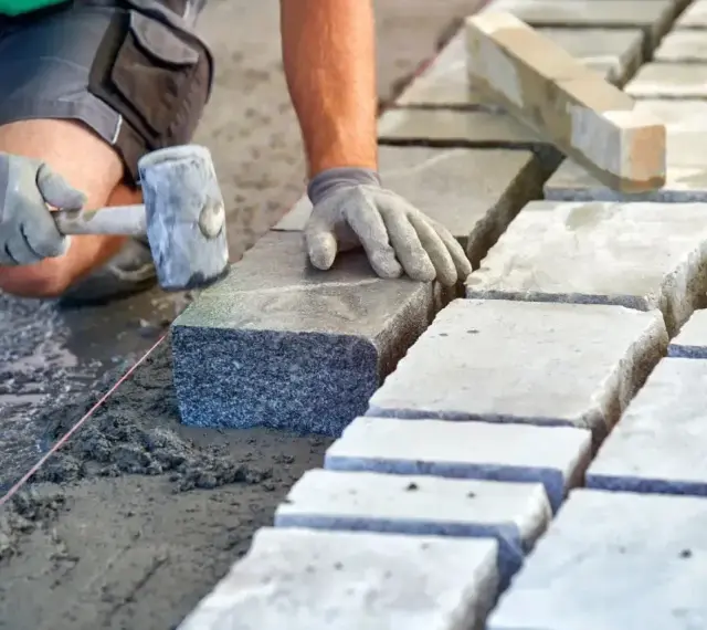 Ręce w rękawiczkach układają kamienne kostki na podsypce. Widać młotek i sznur geodezyjny. Prawidłowe suchy beton proporcje to klucz do trwałej nawierzchni.