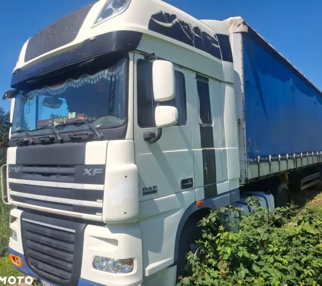 Ciężarówka DAF XF 105 z niebieską plandeką stoi na trawie, częściowo zasłonięta przez krzaki.