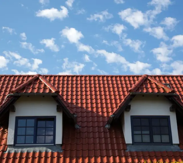 Dachówka ceramiczna w kolorze ceglastym pokrywa dach domu z dwoma lukarnami. Różne rodzaje dachówek tworzą harmonijną całość.
