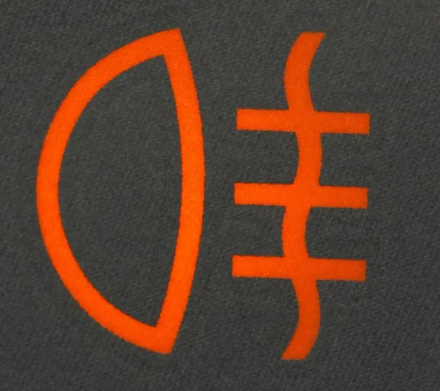Pomarańczowy symbol świateł mijania na ciemnoszarym tle. Ikona przedstawia reflektor z trzema poziomymi liniami.
