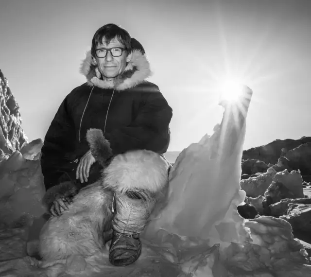 Ein Mann in traditioneller Inuit-Kleidung sitzt in eisiger Landschaft. Die Sonne scheint hell.