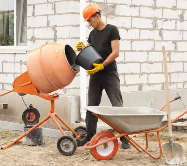 Budowlaniec wsypuje cement do betoniarki 100l. Obok stoi taczka i łopata.