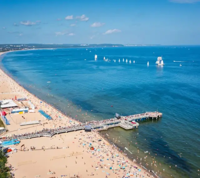 Słoneczna plaża z molo, tłumy ludzi i żaglówki na morzu – idealne miejsce na tanie wycieczki z Gdańska.