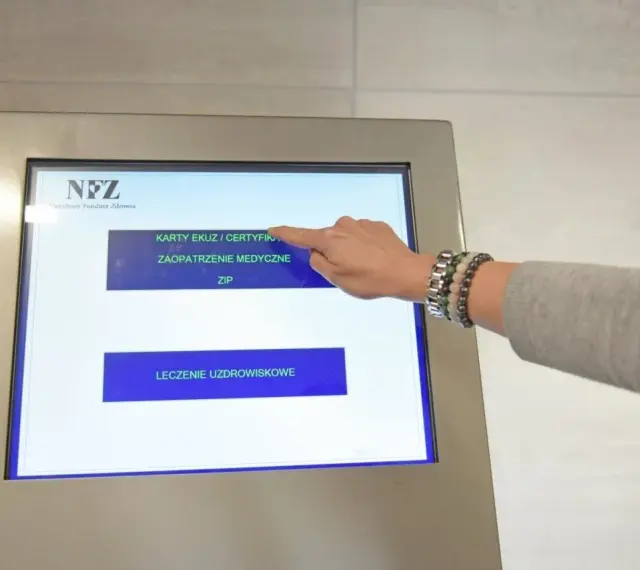 Dłoń dotyka ekranu automatu NFZ, wybierając opcję "Karty EKUZ / Certyfikaty".