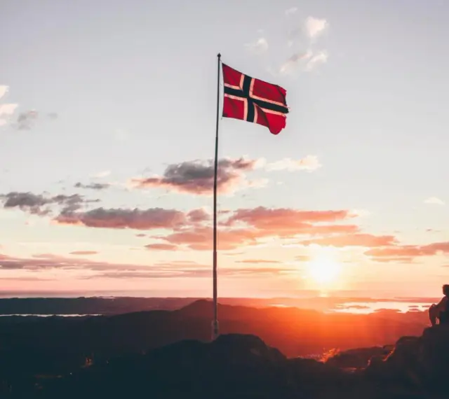 Zachód słońca w Norwegii. Dwie osoby siedzą na skale, patrząc na krajobraz. Flaga Norwegii powiewa na maszcie. Czy do Norwegii można lecieć na dowód osobisty?