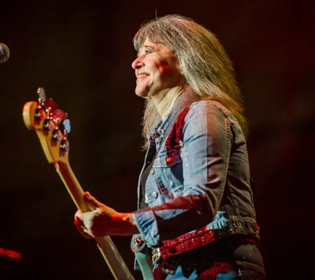 Suzi Quatro dzisiaj na scenie, z basem w ręku, uśmiecha się do publiczności. Rock'n'rollowa legenda w dżinsowej kurtce.