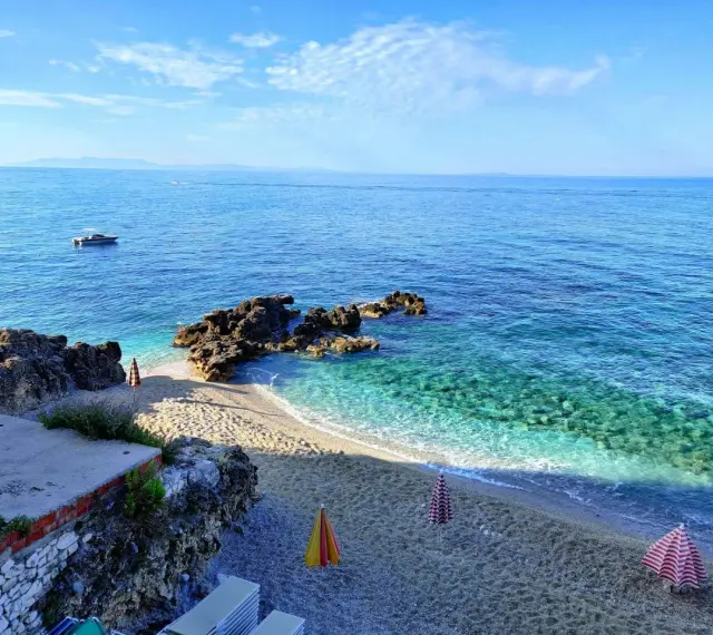 Albania, czerwiec: słoneczna pogoda nad krystalicznie czystym morzem, kamienista plaża z parasolami i łódkami na horyzoncie.
