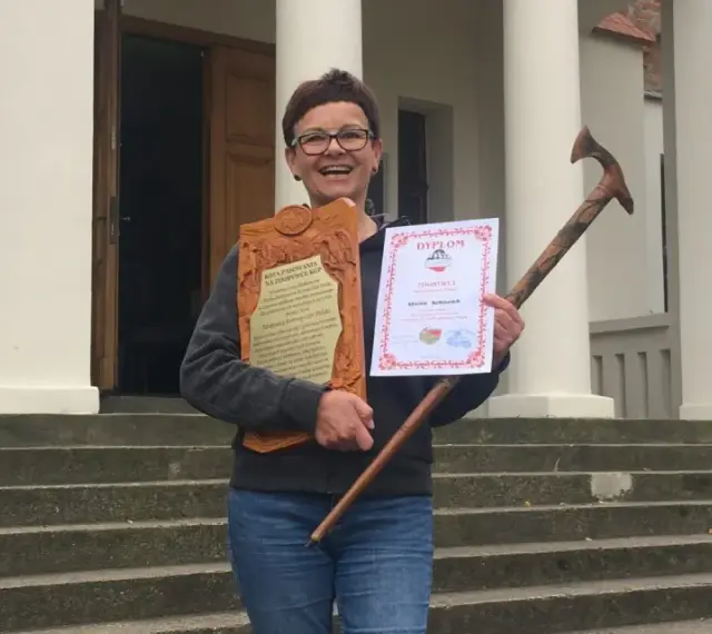 Uśmiechnięta kobieta z dyplomem i drewnianą laską, zdobywczyni Korony Gór Polskich.
