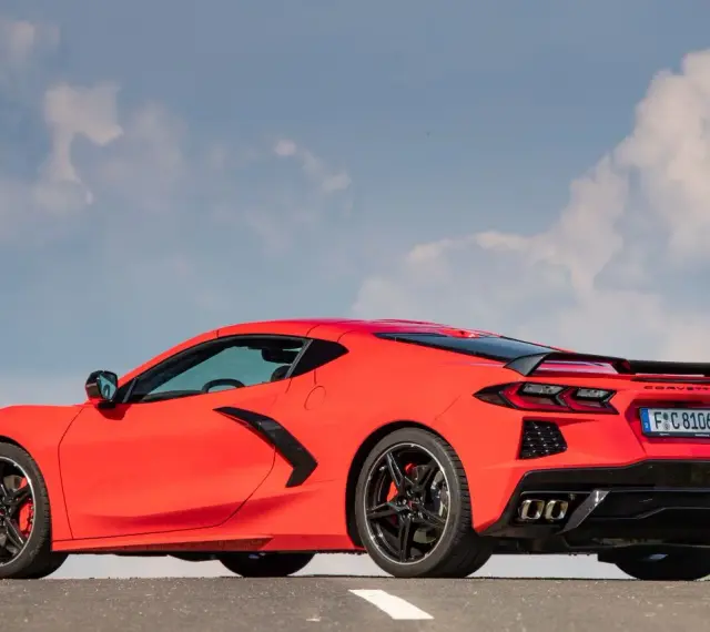 Czerwony Chevrolet Corvette C8 z czarnym spoilerem i felgami, zaparkowany na drodze pod błękitnym niebem z chmurami.