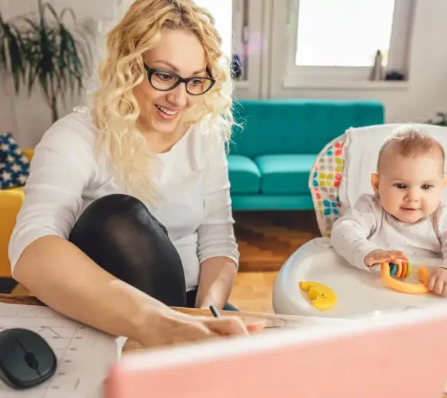 Uśmiechnięta mama pracuje przy laptopie, podczas gdy dziecko bawi się w krzesełku. Planuje zwrot podatku na dziecko.
