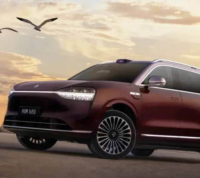 Ciemnoczerwony SUV Aito M9 na tle zachodzącego słońca, z ptakami w locie.