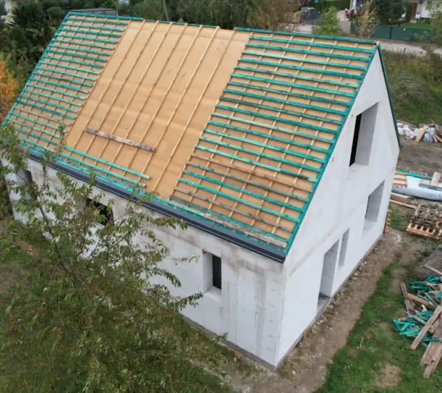 Rozbudowa domu w toku. Dom z białego pustaka, na dachu widoczna więźba i łaty. Wokół materiały budowlane.