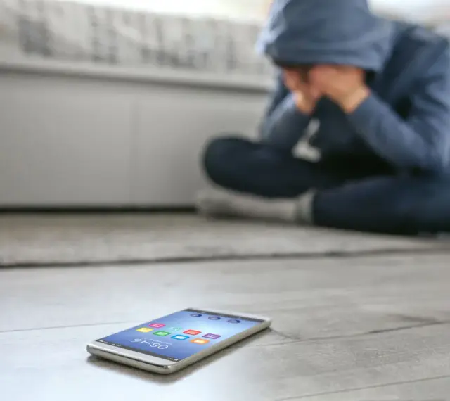 Osoba w kapturze siedzi na podłodze, ukrywając twarz w dłoniach. Obok leży telefon, symbolizujący izolację i dystymię.