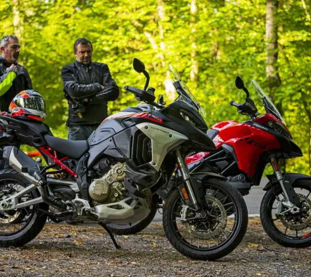 Dwóch motocyklistów z kaskiem na siedzeniu obok dwóch motocykli Ducati Multistrada, szary i czerwony, na tle leśnej drogi.