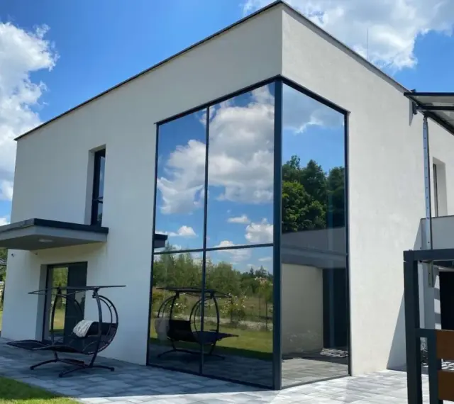 Nowoczesny dom z imponującym, narożnym oknem weneckim, w którym odbija się błękitne niebo z chmurami i zieleń drzew.