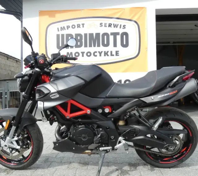 Czarny motocykl Aprilia Shiver 900 z czerwonym stelażem, zaparkowany przed salonem motocyklowym.