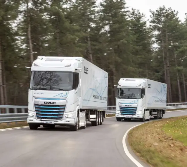 Dwa białe elektryczne DAF XG Plus z naczepami jadące po krętej drodze wśród drzew.