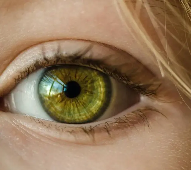 Zielona soczewka oka z refleksami światła, otoczona jasną skórą i blond włosami.