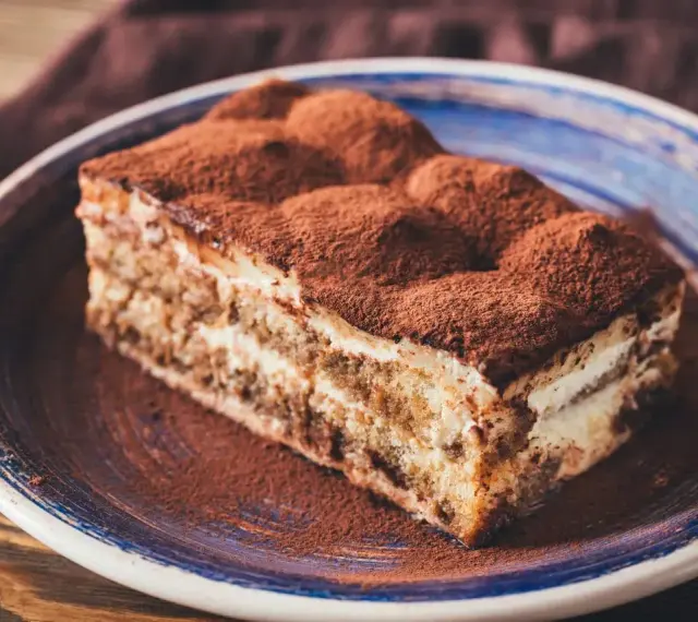 Pyszny kawałek tiramisu z mascarpone, posypany kakao, na niebieskim talerzu. Idealny na deser.