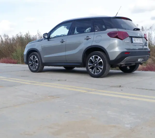 Szary Suzuki Vitara Hybrid z czarnym dachem stoi na betonowej nawierzchni z żółtymi pasami.