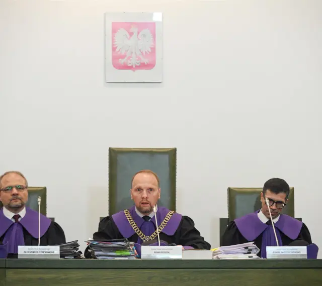 Trzech sędziów w togach podczas jawnego posiedzenia sądu. Na ścianie wisi godło Polski.