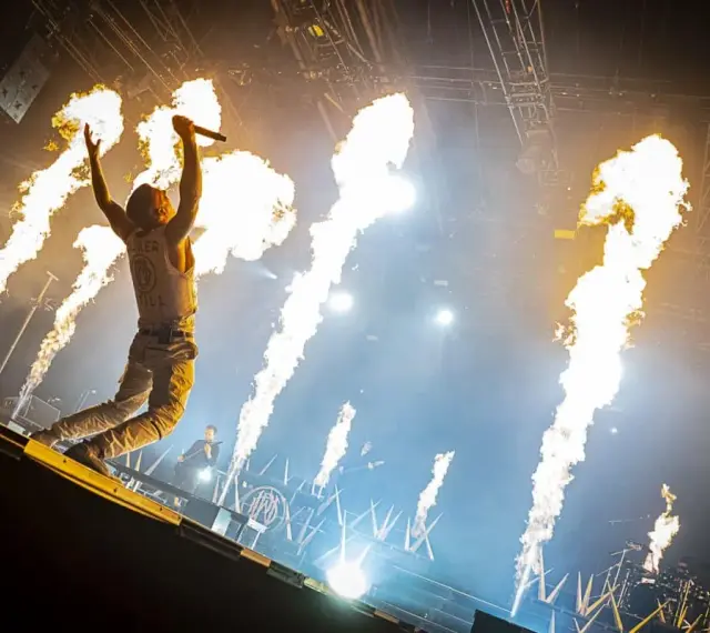 Un chanteur de groupe hard rock des années 2000, micro en main, saute sur scène entouré de jets de flammes spectaculaires.