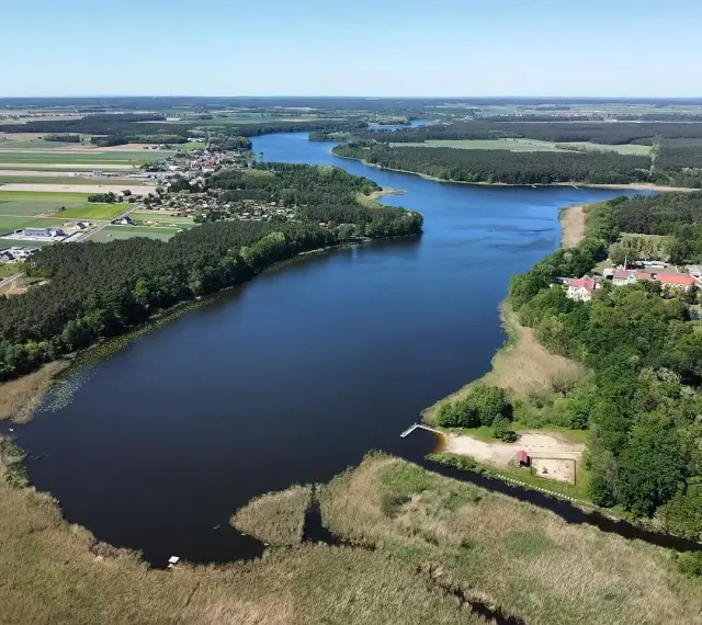 Malownicze jezioro Wojnowskie otoczone lasami i polami. W oddali widać zabudowania wsi, a na brzegu plażę z pomostem.
