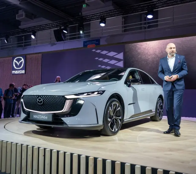 Elegancki, szary sedan Mazda6e prezentowany na scenie przez mężczyznę w garniturze.