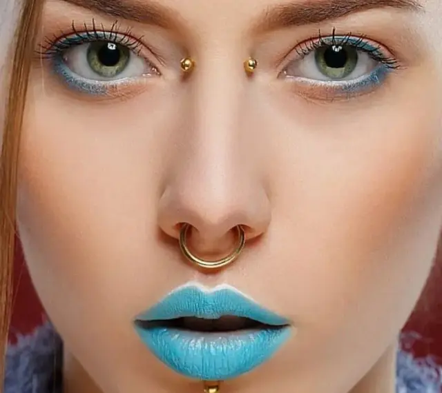 Kobieta z błękitnymi ustami i złotym septum. Jej oczy podkreślone są niebieskim cieniem, a między brwiami widoczny jest bridge piercing.