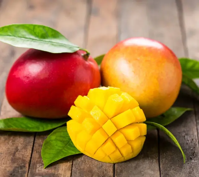 Dwa soczyste mango, jedno czerwone, drugie pomarańczowe, leżą na drewnianym blacie. Obok nich pokrojone w kostkę, złociste wnętrze owocu. To przypomina o tropikalnych krajach, skąd pochodzi mango.