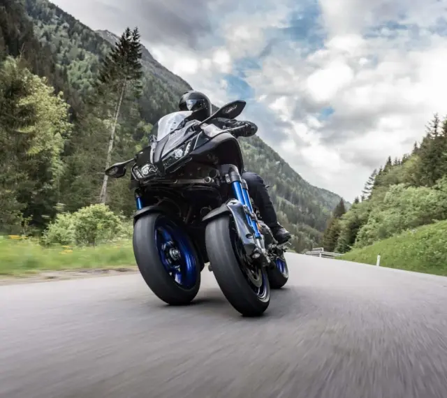 Motocyklista na Yamaha Niken pokonuje górską drogę, otoczony zielonymi lasami i skalistymi szczytami.