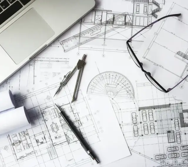 Na biurku architekta leżą plany budowlane, laptop, okulary i cyrkiel. Trwają prace nad przedmiarem robót.
