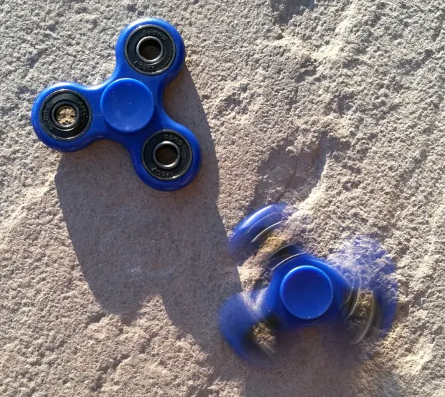 Dwa niebieskie fidget spinnery na piasku, jeden nieruchomy, drugi w ruchu, przypominają czasy, kiedy fidget spinnery były popularne.