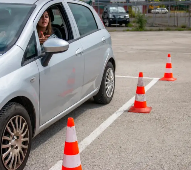 Młoda kobieta za kierownicą srebrnego samochodu ćwiczy jazdę na placu manewrowym. To może być jej droga do zdobycia prawa jazdy 17 lat.
