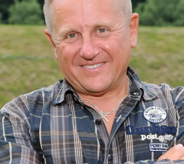 Uśmiechnięty mężczyzna w kratę, z krzyżykiem na szyi. To Bercik, znany z serialu.