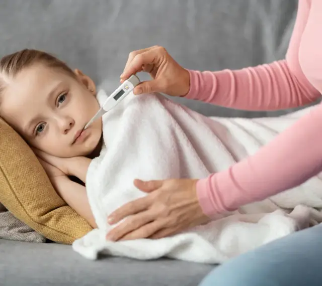 Chore dziecko z gorączką może iść na dwór? Mama mierzy temperaturę chorej córce leżącej pod kocem.